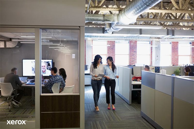 Due donne camminano in un moderno ufficio open space, impegnate in una conversazione e circondate da postazioni di lavoro.