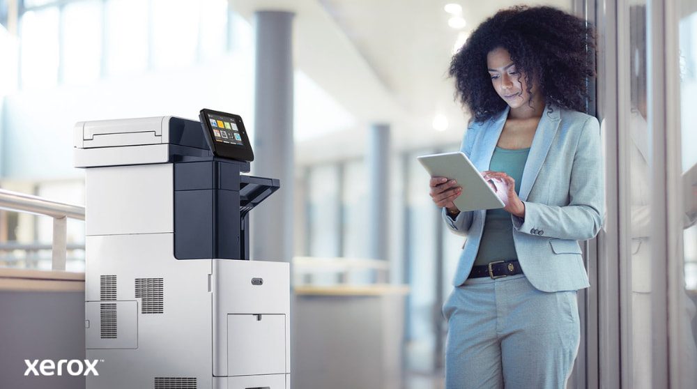 Xerox® ConnectKey®: connettività mobile Una persona è in piedi accanto a una stampante multifunzione Xerox VersaLink B605, osserva e interagisce con un dispositivo tablet, illustrando come ConnectKey consente l'interazione mobile con la stampante.