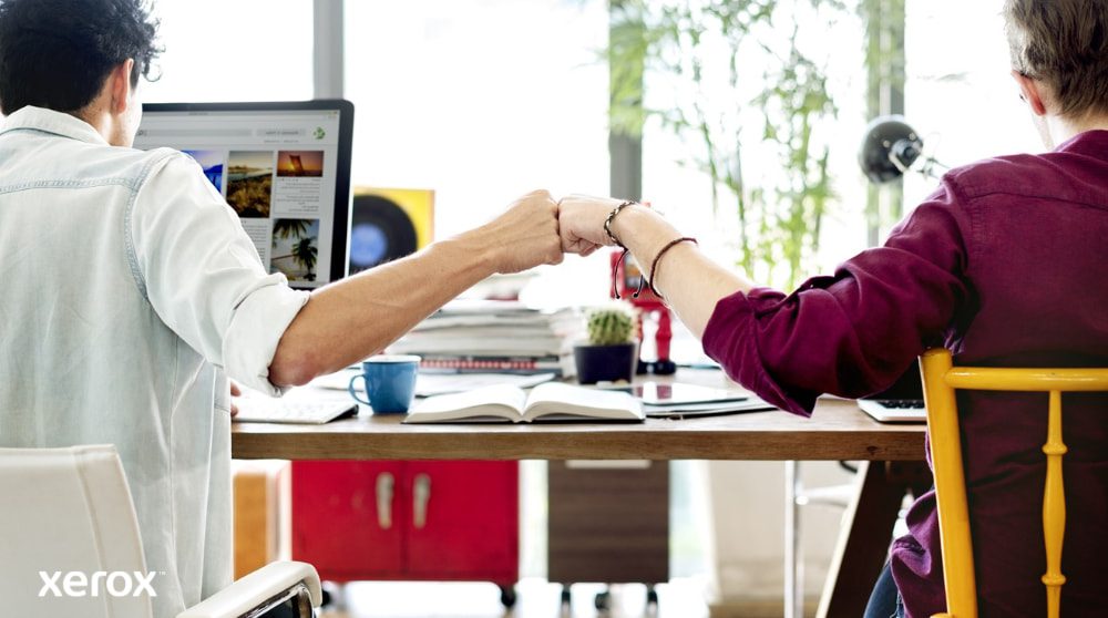 Xerox® Smart Start: lavoro di squadra e collaborazione Due persone sedute a una scrivania, con le spalle rivolte alla telecamera, si salutano con un pugno, simbolo di lavoro di squadra e collaborazione di successo. Una di loro guarda lo schermo di un computer.